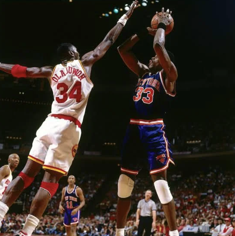 Two basketball players in action during a game. One player, in a white jersey with number 34, is attempting to block a jump shot from another player in a blue jersey with number 33. A crowd is visible in the background.