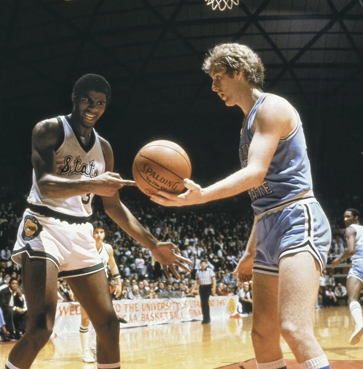 Two basketball players on a court during a game. One player in a white uniform holds a basketball, while the other in a light blue uniform reaches for it. The background shows spectators and a large scoreboard.