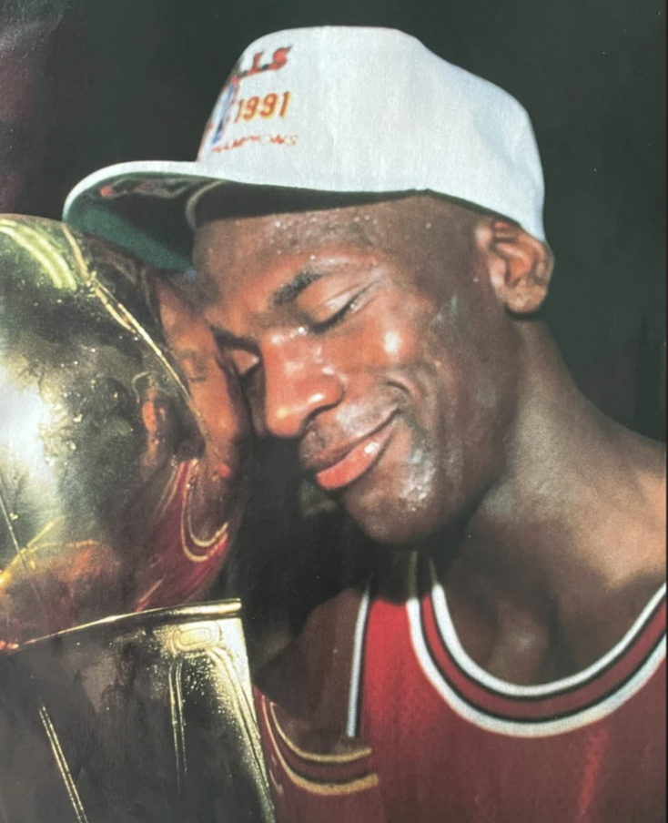 A basketball player in a red jersey and white cap with "1991" printed on it smiles and presses his face against a shiny gold trophy. The scene captures a moment of joy and triumph.