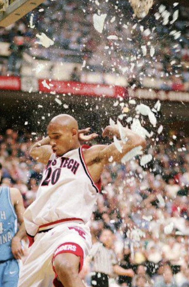 Basketball player wearing a Texas Tech jersey shatters a backboard while dunking. Glass fragments surround him as he hangs onto the rim. The court and spectators are blurred in the background.