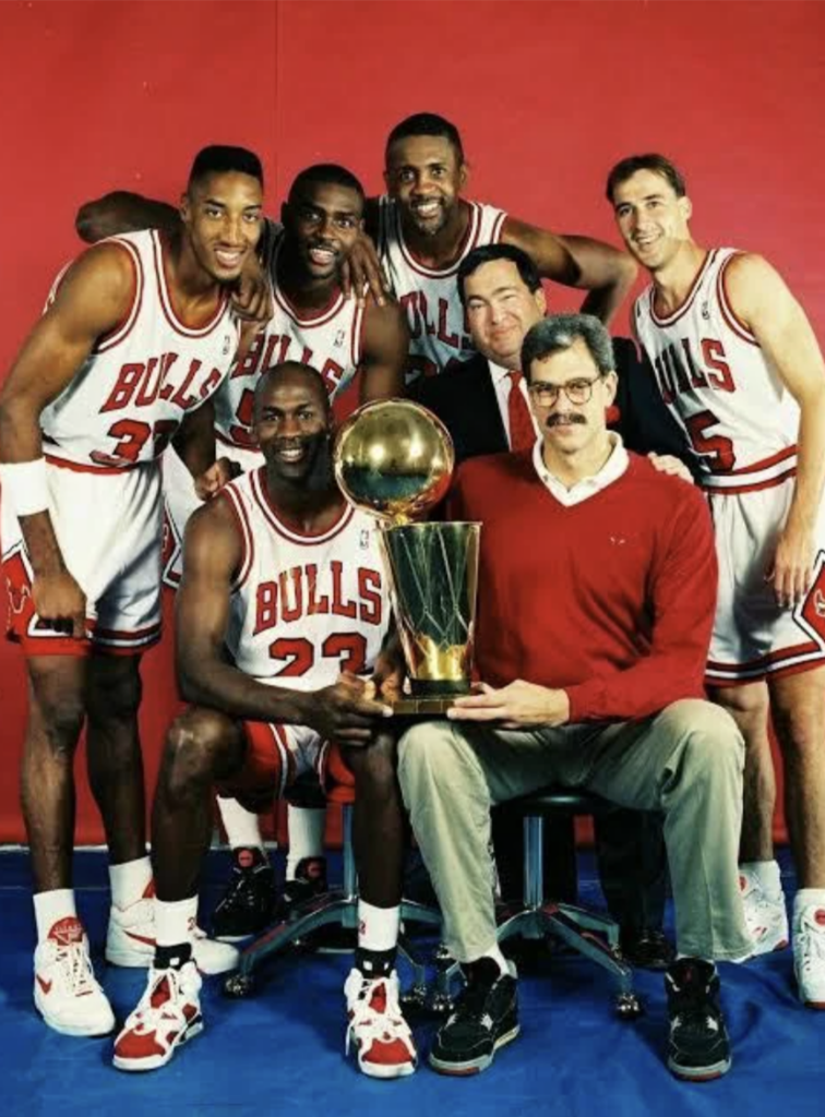 A group of basketball players and staff in Chicago Bulls uniforms pose with a large trophy against a red background. Two of them are kneeling, holding the trophy, while the others stand behind them, smiling.