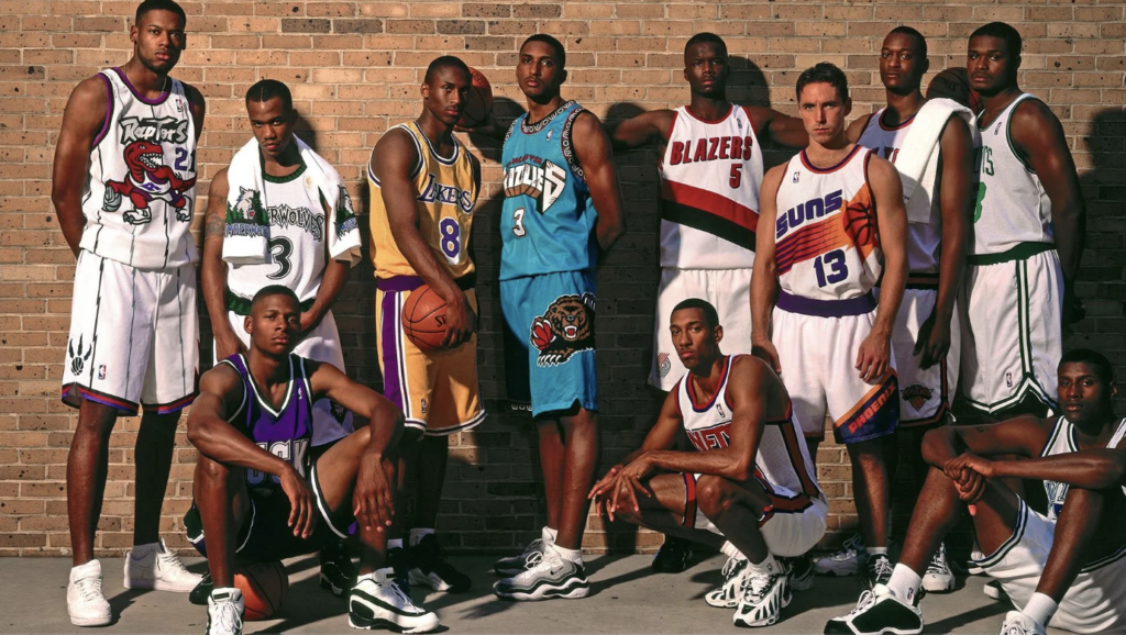 A group of basketball players wearing 1990s NBA jerseys pose against a brick wall. The jerseys feature teams like the Raptors, Lakers, Timberwolves, Grizzlies, Trail Blazers, Suns, and Bucks. The players are standing and kneeling in various poses.