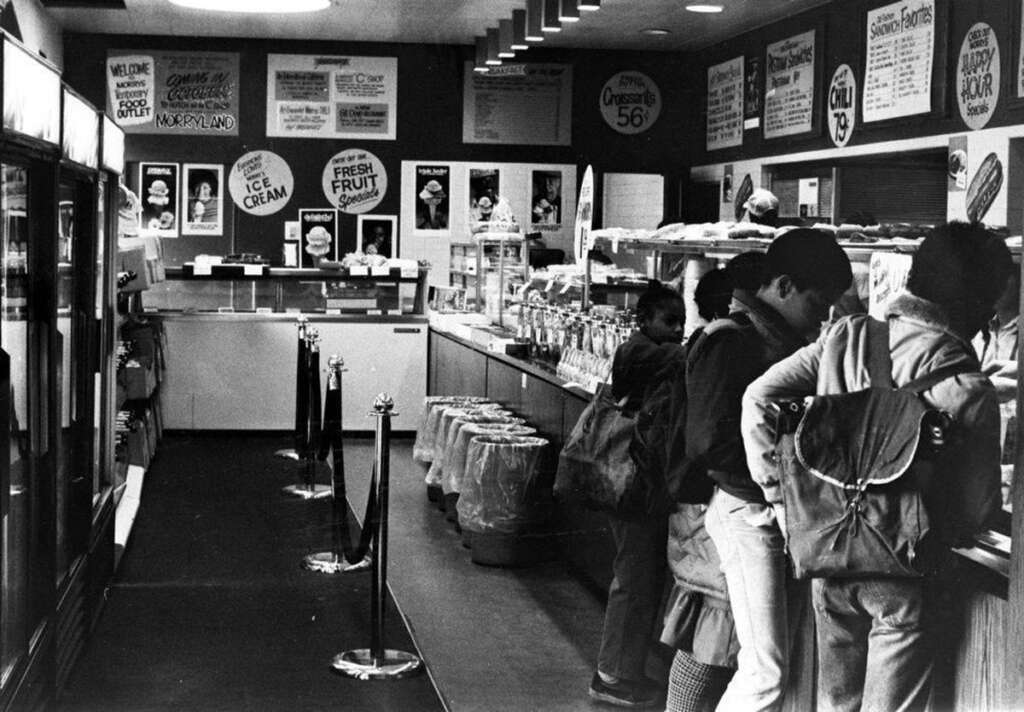 Black and white photo of people standing in line at an ice cream shop. The interior has various signs advertising flavors and prices. The countertop displays toppings, and there are ropes to guide the line. The atmosphere is busy and casual.