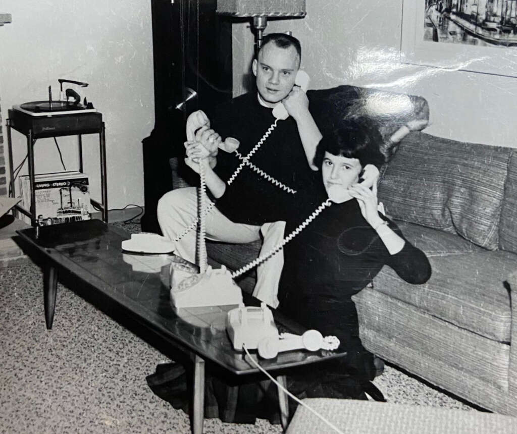 A black-and-white photo of a man and a woman sitting in a living room on a couch. Both are talking on old-fashioned rotary phones with long cords. A coffee table is in front of them, and a record player is visible in the background.