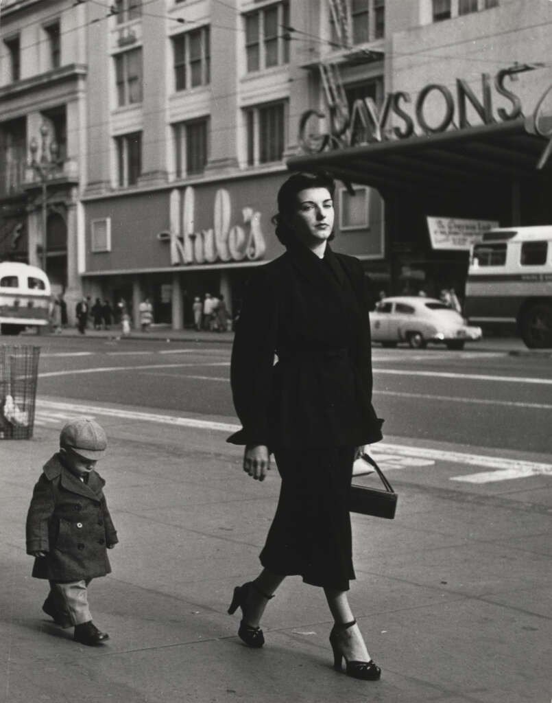 A woman in a dark coat and heels walks on a city street, holding a purse. A small child in a coat and hat follows her. Cars and storefronts, including "Mule's" and "Gaysons," are visible in the background. The scene has a vintage feel.