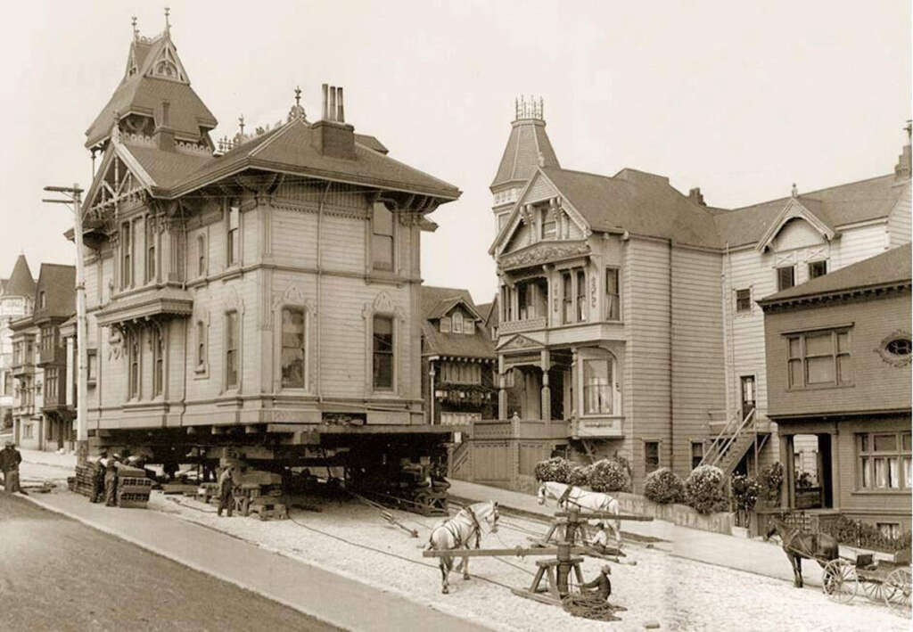 A historic Victorian-style house is being relocated down a street using horse-drawn equipment. The house is elevated on wooden beams with nearby similarly-styled buildings lining the street.