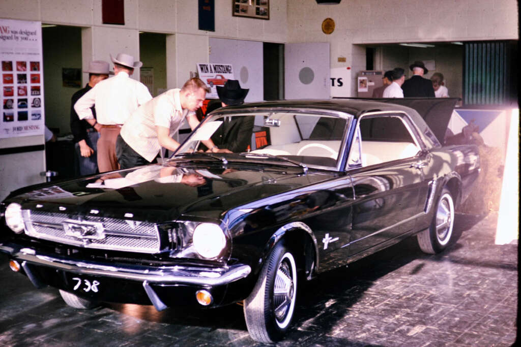 A vintage black Ford Mustang is displayed indoors. A man leans over to inspect the interior. Several people in 1960s attire stand nearby, some wearing hats. The background shows posters and a door.