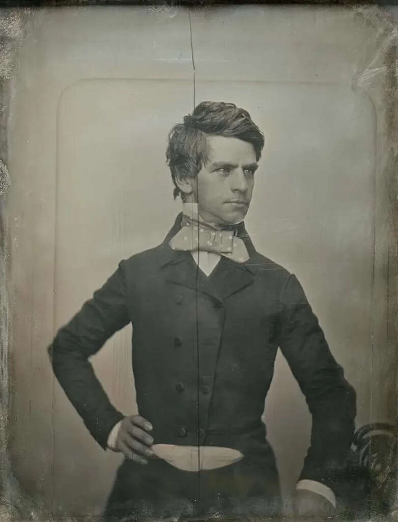 A vintage daguerreotype portrait of a young man with short hair, posing with one hand on his hip. He's wearing a double-breasted suit and a large bow tie. The image has a vertical crease down the center and exhibits signs of age.