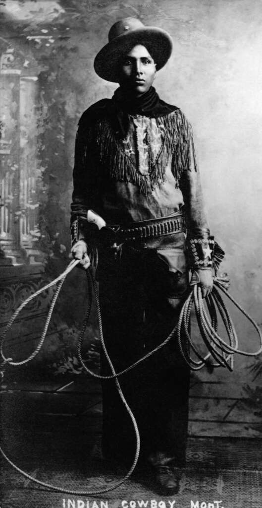 Black and white photo of a cowboy wearing a hat and fringed jacket, holding a lasso. A pistol is visible at his waist. The background is a vintage-style painted backdrop. The text at the bottom reads "Indian Cowboy - Moet.