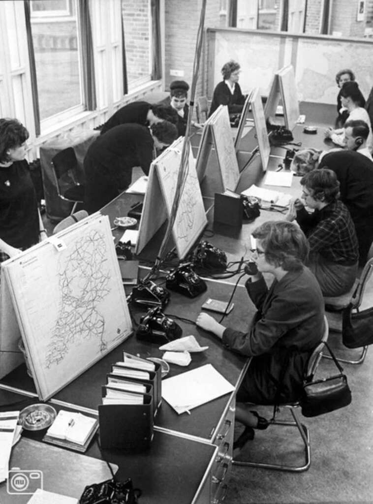 Black and white image of a busy office with women working at a long desk. They are using maps on wooden boards and telephones. Papers and folders are scattered on the desks. Large windows and maps on the wall are visible in the background.