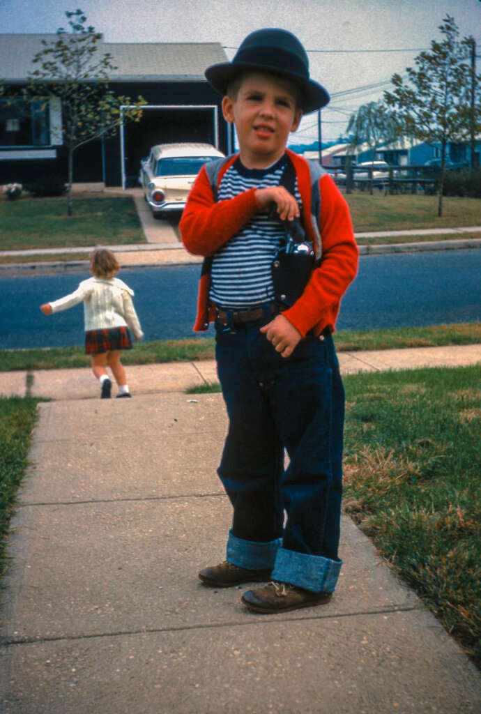 A young boy wearing a bowler hat, red cardigan, striped shirt, and jeans stands on a sidewalk, holding a camera. In the background, a girl in a plaid skirt walks away. A vintage car and house are visible in the distance.