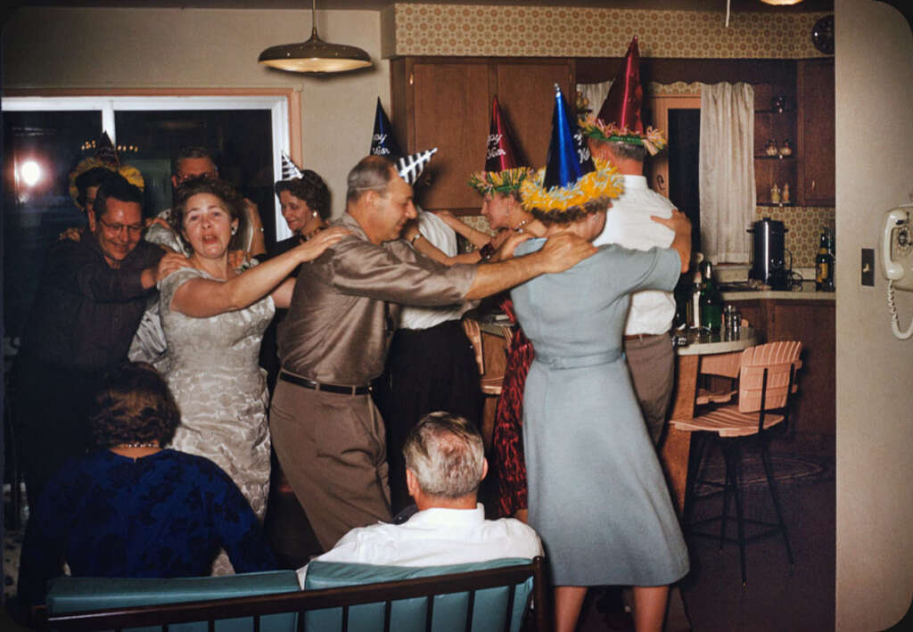 People in a living room having a party. They are wearing colorful party hats and dancing in a conga line. The room is decorated with mid-century furniture and has a cozy atmosphere. Some guests are seated, enjoying the festivities.