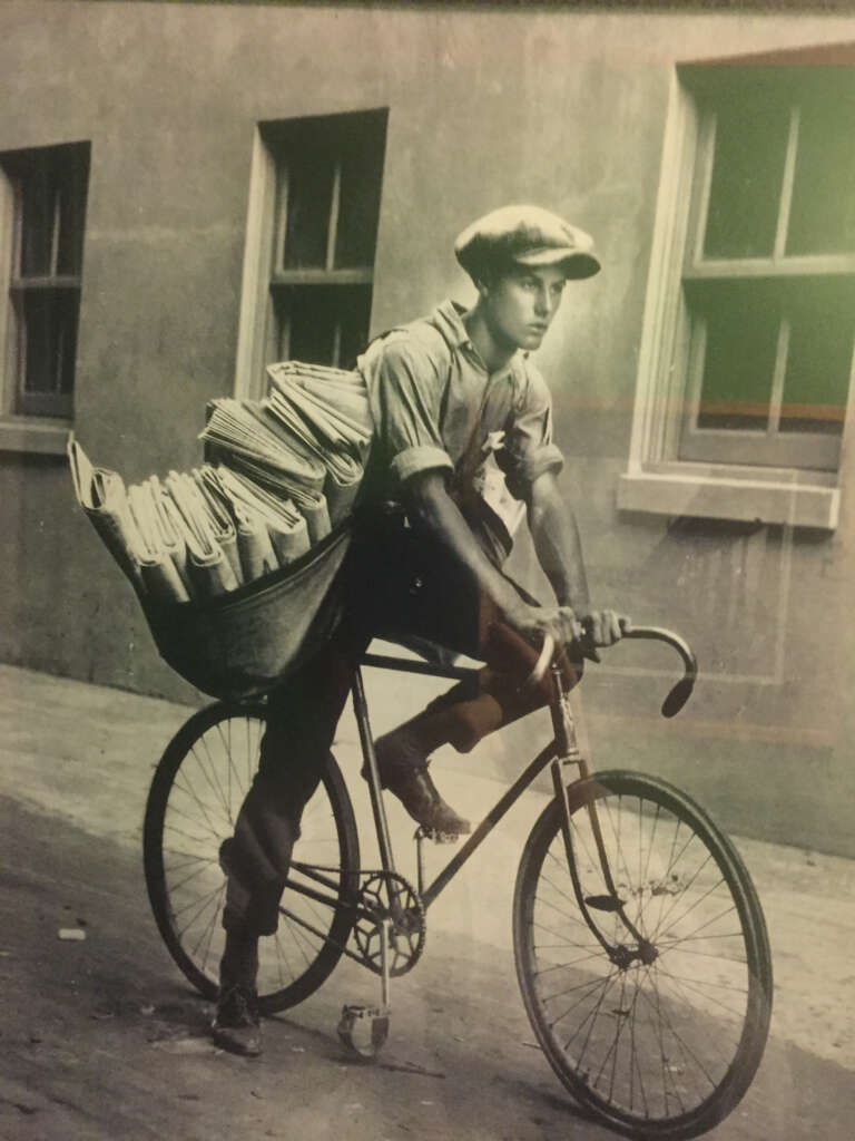 A vintage photograph of a young newspaper delivery boy riding a bicycle. He wears a cap and carries a large sack full of newspapers on the bike. The background shows a building with two windows.