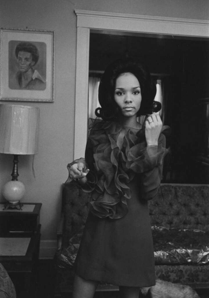 A woman with long hair stands in a living room, wearing a dress with ruffled details. She holds a small object. Behind her, there's a sofa, a table with a lamp, and a portrait on the wall. The room has a classic, vintage feel.