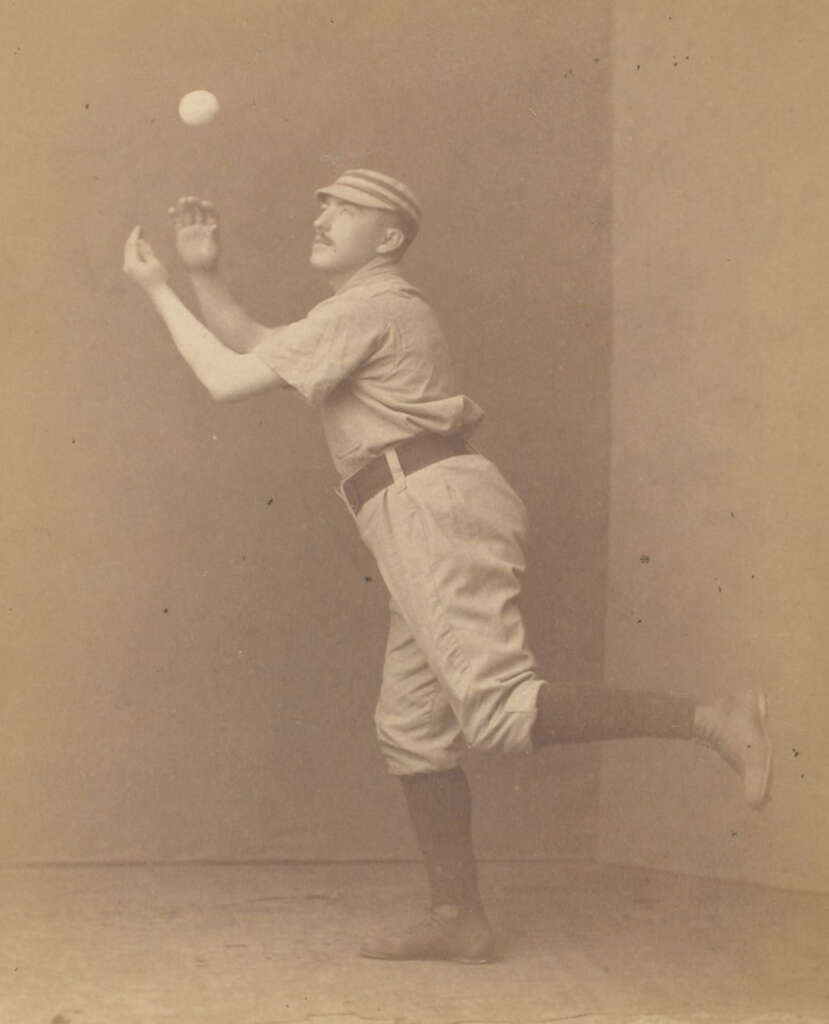 A vintage sepia-toned photograph of a baseball player in mid-action. He's dressed in early baseball attire, including a cap, and is poised to catch a ball. The player is standing on one foot, with a slightly blurred ball above him.