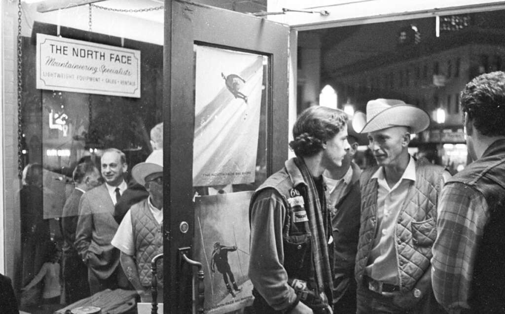 A black-and-white photo of people, some wearing cowboy hats and quilted vests, standing in and around a store entrance. The store, The North Face, displays skiing posters on the door and window. Streets and buildings are visible outside at night.