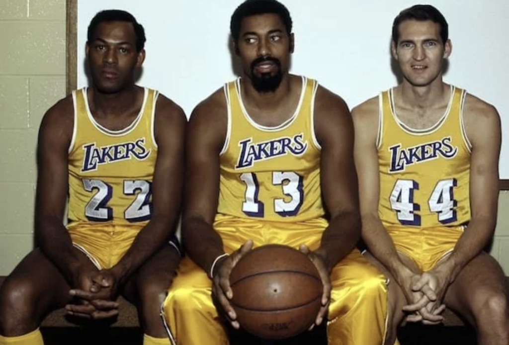 Three basketball players in yellow and blue Lakers uniforms are sitting on a bench. The player in the center holds a basketball. They are in a locker room with a pale green wall in the background.