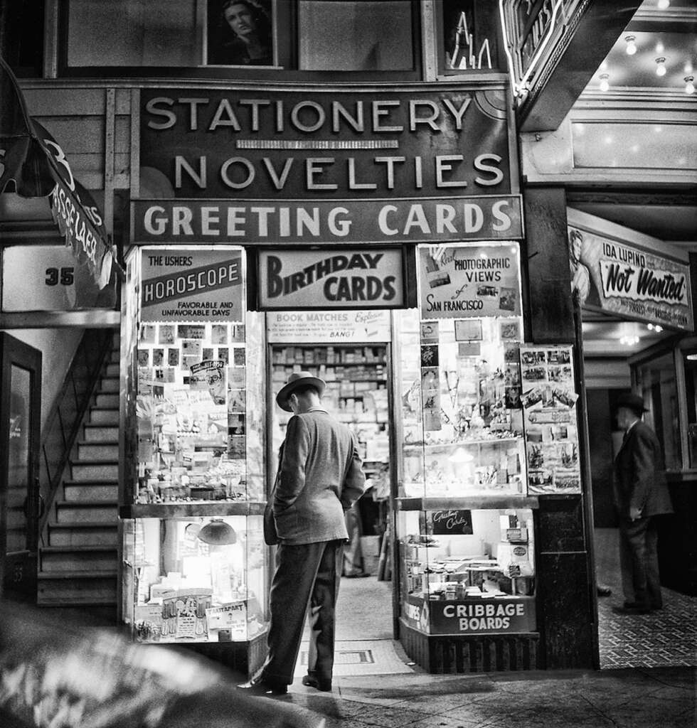 A man in a suit and hat stands outside a vintage stationery and novelties shop. The storefront displays signs for horoscopes, greeting cards, photographic views, and more. The shop is brightly lit, showcasing a variety of items through the window.