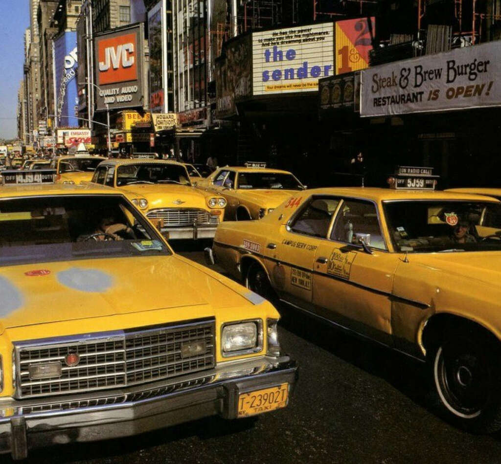 A busy street scene with yellow taxis filling the road in front of various businesses, including a Steak & Brew Burger restaurant with a sign saying "Restaurant is Open." Billboards and signs are visible in the background.