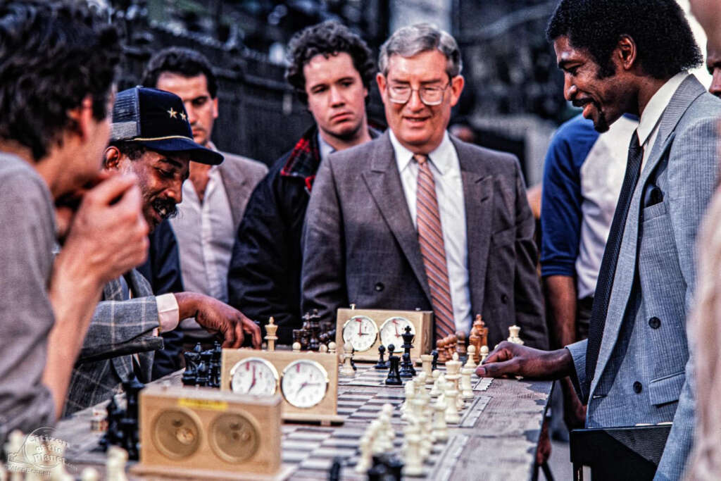 A group of men gathers around an outdoor chess game. Two players are focused on the board, one man points at a piece. Observers, including a man in a suit and tie, watch intently. Two chess clocks are positioned on the table.