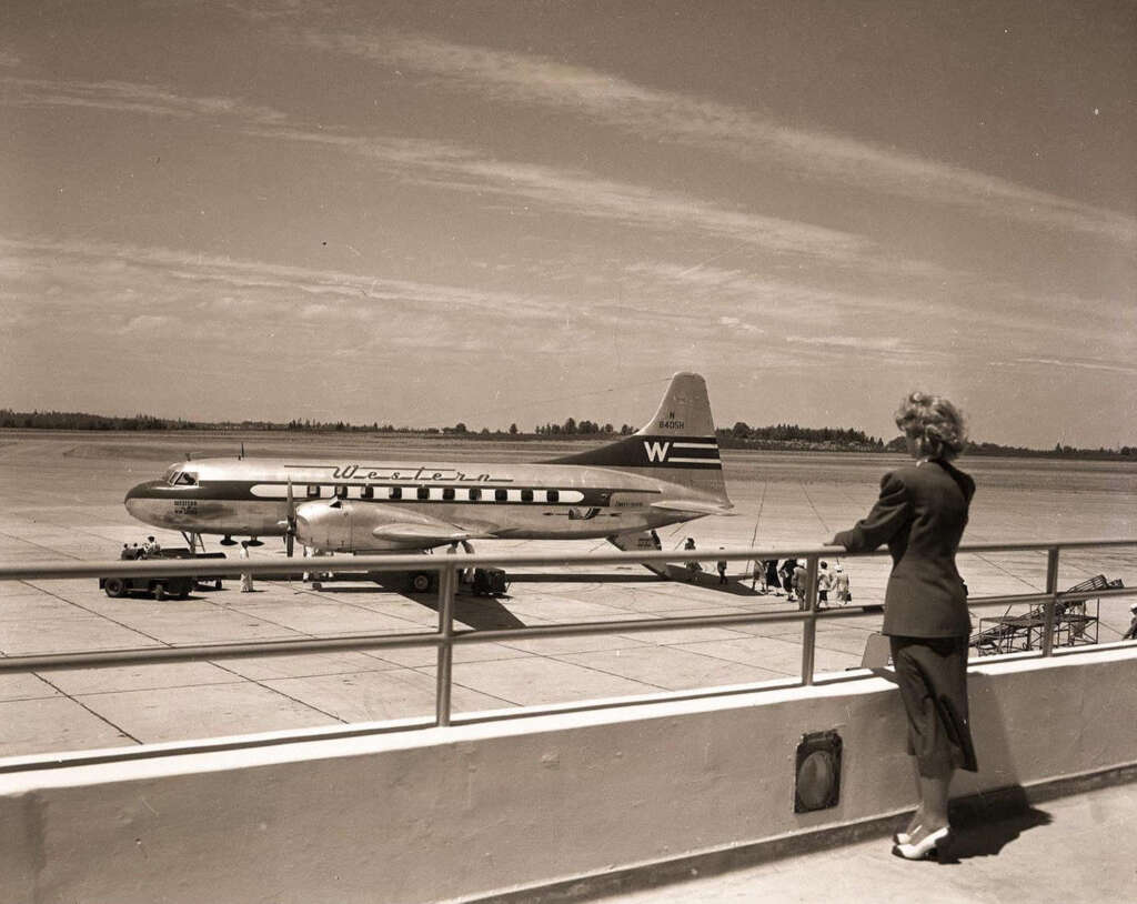 A woman in a suit stands at an airport viewing area, looking at a vintage passenger airplane on the tarmac. The aircraft has "Western" written on the fuselage. A few people are near the plane. The sky is partly cloudy.