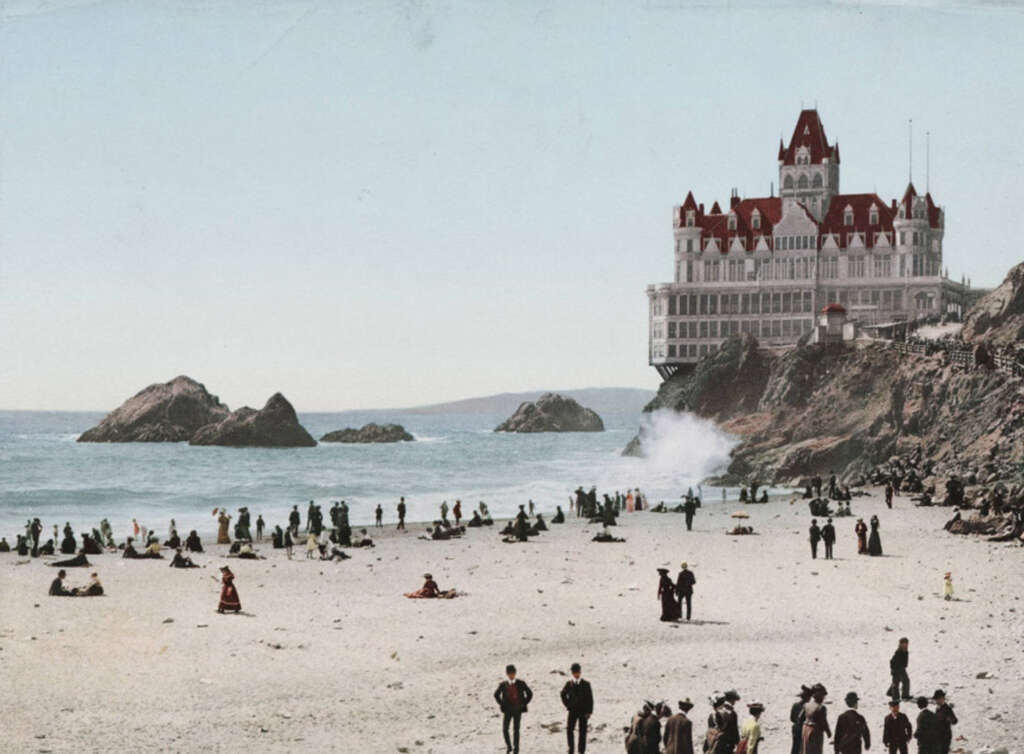 A Victorian-style building with red roofs overlooks a beach crowded with people dressed in vintage clothing. Waves crash against rocks in the ocean, and the sky is clear.