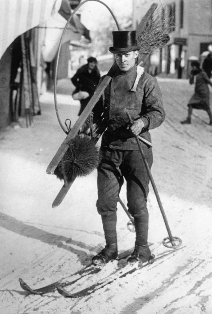 A vintage photograph shows a man in a top hat standing on skis in a snowy street. He carries skis, a circular hoop, and brushes slung over his shoulder. Another person is in motion in the background. The scene appears to be from an earlier era.