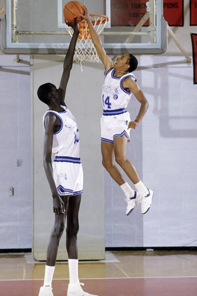 Two basketball players in uniforms are on a court near a hoop. The taller player attempts to block the shot with one hand, while the shorter player, wearing number 14, jumps high with the ball, reaching towards the basket.