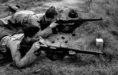 Two people lie on the ground in a grassy area, aiming rifles. They are on their stomachs, focused on their targets. One person has a hat on the ground nearby. The setting suggests a target practice or military exercise.