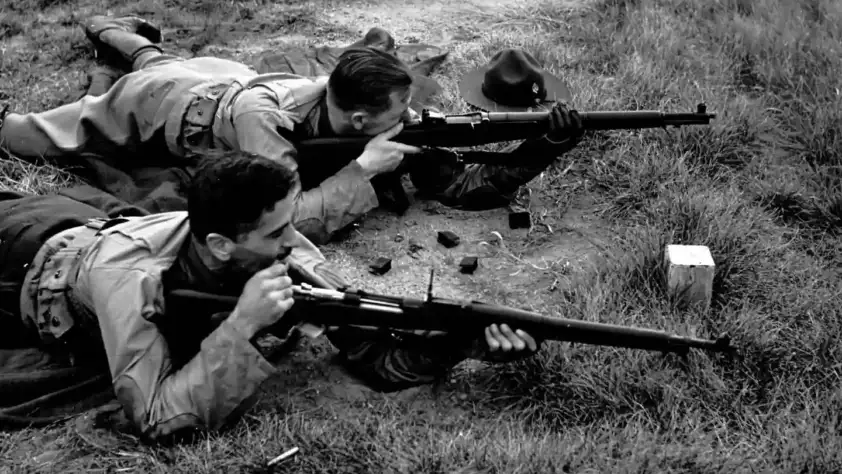 Two people lie on the ground in a grassy area, aiming rifles. They are on their stomachs, focused on their targets. One person has a hat on the ground nearby. The setting suggests a target practice or military exercise.