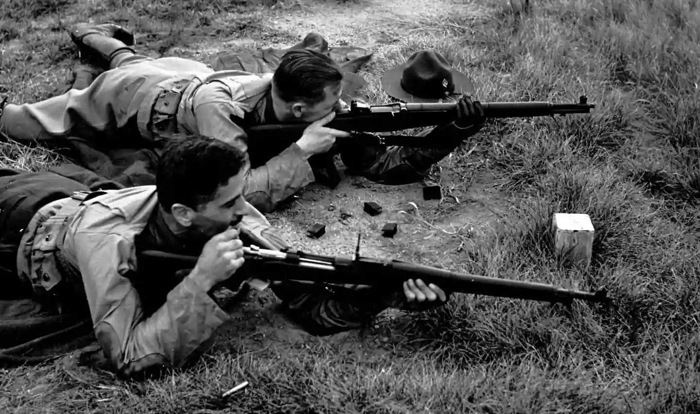 Two people lie on the ground in a grassy area, aiming rifles. They are on their stomachs, focused on their targets. One person has a hat on the ground nearby. The setting suggests a target practice or military exercise.