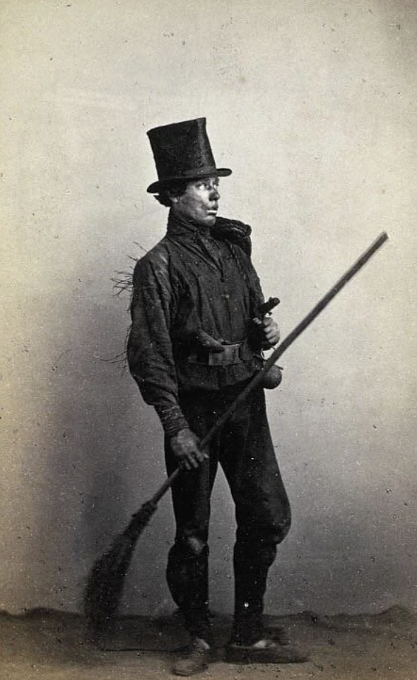 A Victorian-era chimney sweep stands holding a brush and pole. He wears a tall top hat, a worn jacket, and trousers. The image is black and white, highlighting the historical attire and profession.