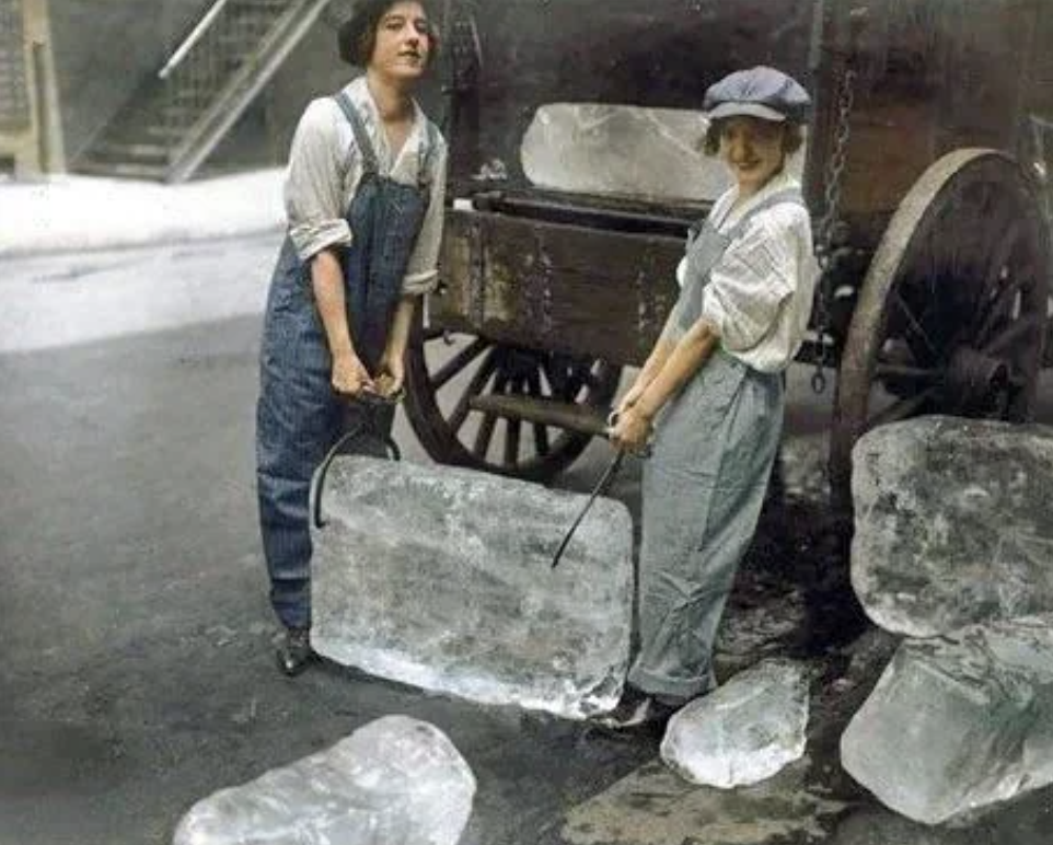 Two people in overalls and caps carry a large block of ice using hooks in front of a wagon with more ice blocks. The setting appears to be a historical urban street scene.