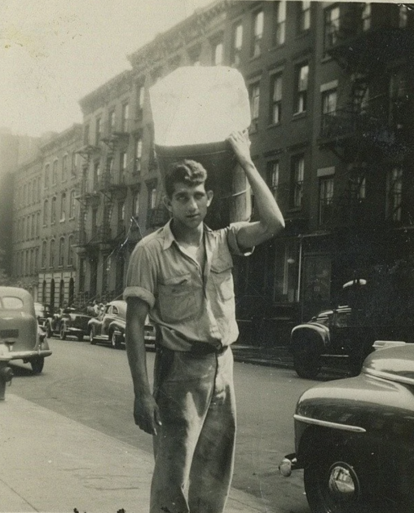 A black-and-white photo of a young man carrying a large box on his head. He is wearing a button-up shirt and pants. The street is lined with cars and brick buildings in the background. The scene appears to be from a past era.