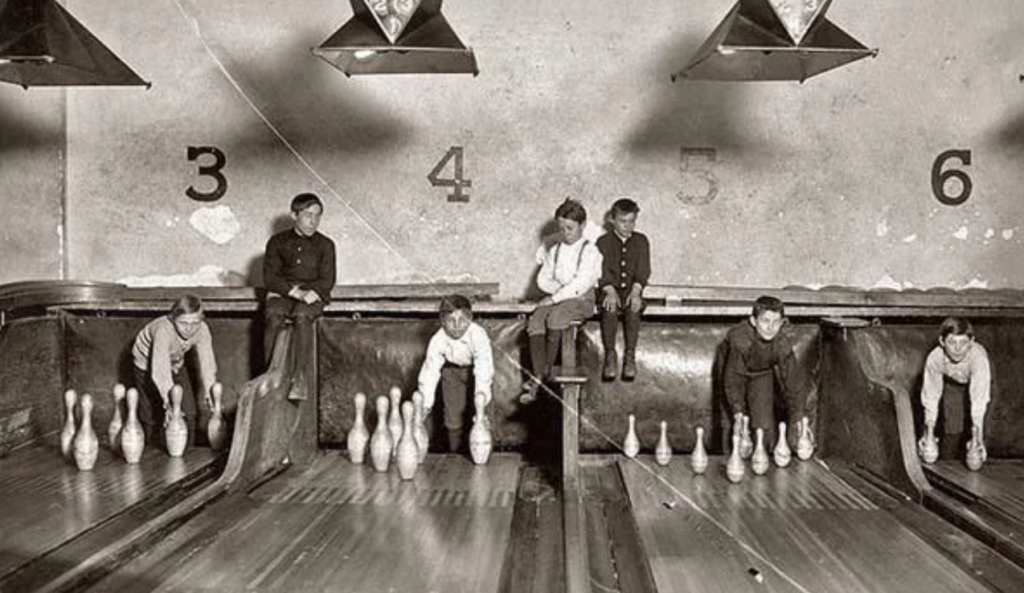 A vintage black-and-white image of boys manually setting up bowling pins in an old-fashioned bowling alley. The boys sit or kneel on raised platforms behind the lanes, while numbers 3 to 6 are visible on the wall above them.