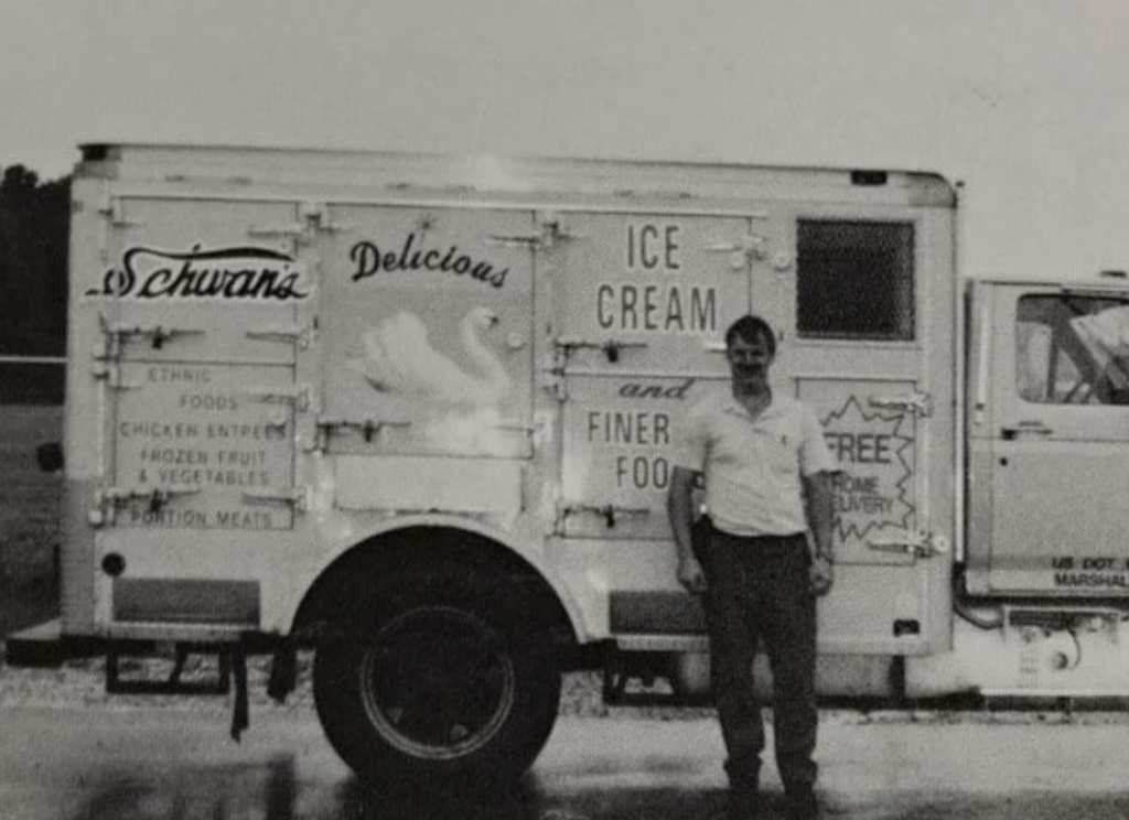 A man stands in front of a vintage delivery truck with "Schwann's Delicious Ice Cream and Finer Foods" written on the side. The truck's design includes a swan illustration and various product advertisements. The man wears a light-colored shirt.