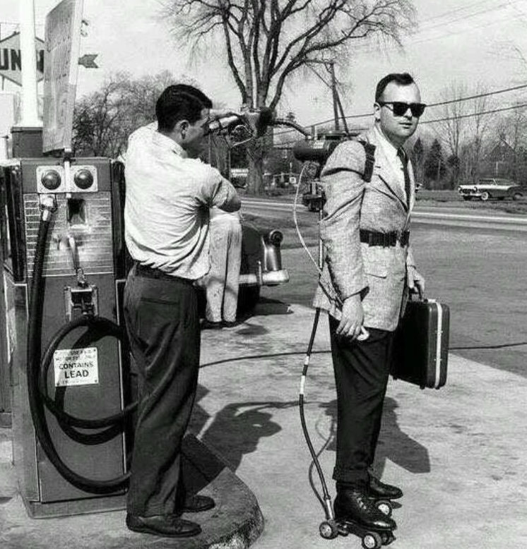 Man in a suit wearing a jetpack and roller skates is being refueled at a gas station by an attendant. He holds a briefcase, and there's a vintage gasoline pump in the background. The setting appears to be from the mid-20th century.