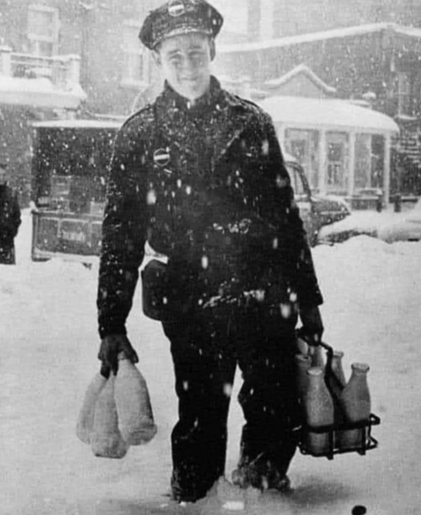 A milkman in a uniform walks through deep snow carrying milk bottles and a bag on a snowy street. A vintage vehicle and houses are visible in the background, capturing a winter scene.