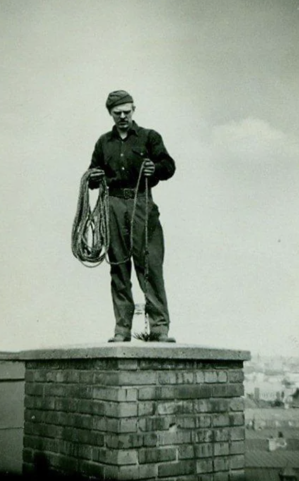 A person wearing a beret and glasses stands on top of a brick chimney, holding a coiled rope. The background shows a cloudy sky and cityscape. The image is in black and white.