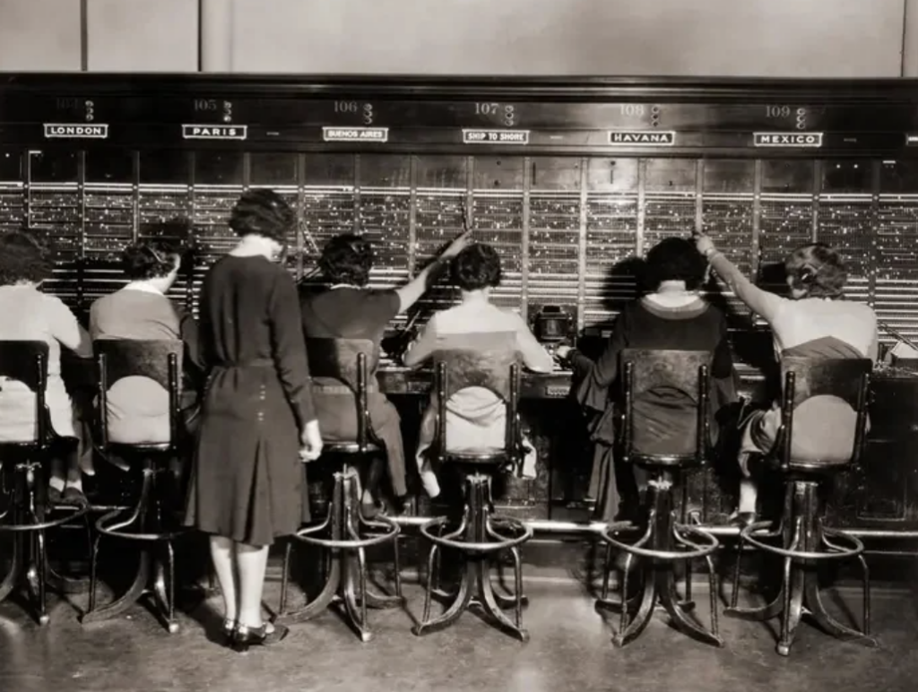 A vintage black-and-white photo shows women operating a large telephone switchboard. They sit on stools, each managing different connections labeled with cities like London, Paris, Havana, and Mexico. A standing woman supervises their work.