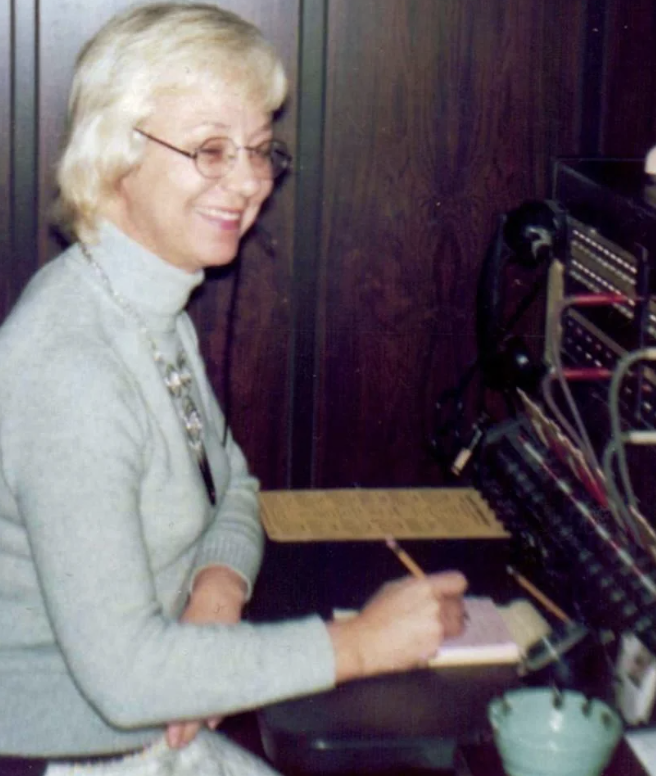 An older woman with short blond hair and glasses is seated at a vintage switchboard. She is wearing a grey turtleneck sweater and smiling while writing on a notepad. The switchboard has numerous wires and plugs.