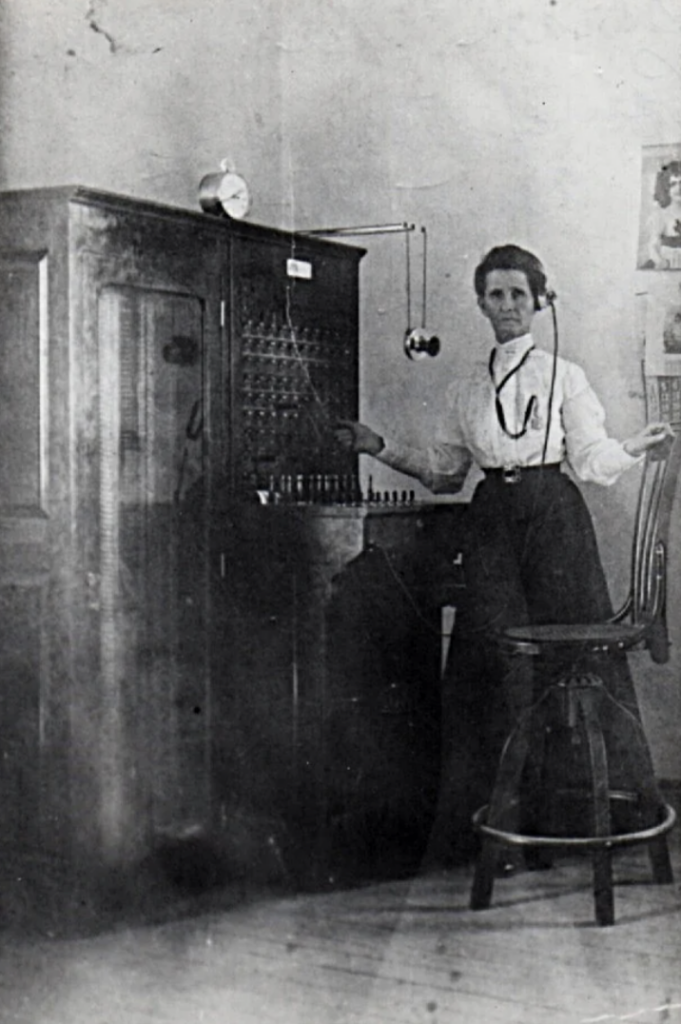 A woman in early 20th-century attire stands next to a vintage telephone switchboard, wearing a headset. She's in a room with minimal decor, and a wooden chair is beside her. A clock and a wall poster are visible in the background.