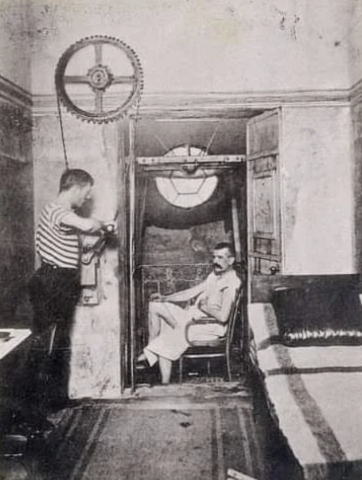 A black-and-white photo showing two men in a room with machinery. One man is seated inside a chamber with a round window, wearing a towel. The other stands outside, operating a mechanism with large gears and chain on the wall.