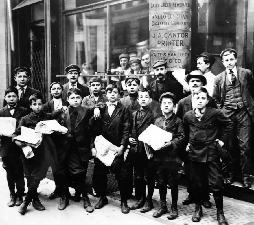 A group of young newspaper boys in early 20th-century attire pose in front of a building. Some hold newspapers, and a few men stand behind them. The scene captures the bustling atmosphere of the era.