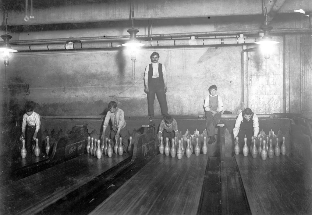 Five people in an old bowling alley setting up pins manually on three lanes. Dimly lit with visible pipes and industrial fixtures on the ceiling. One person stands on a lane, while others crouch near the pins.