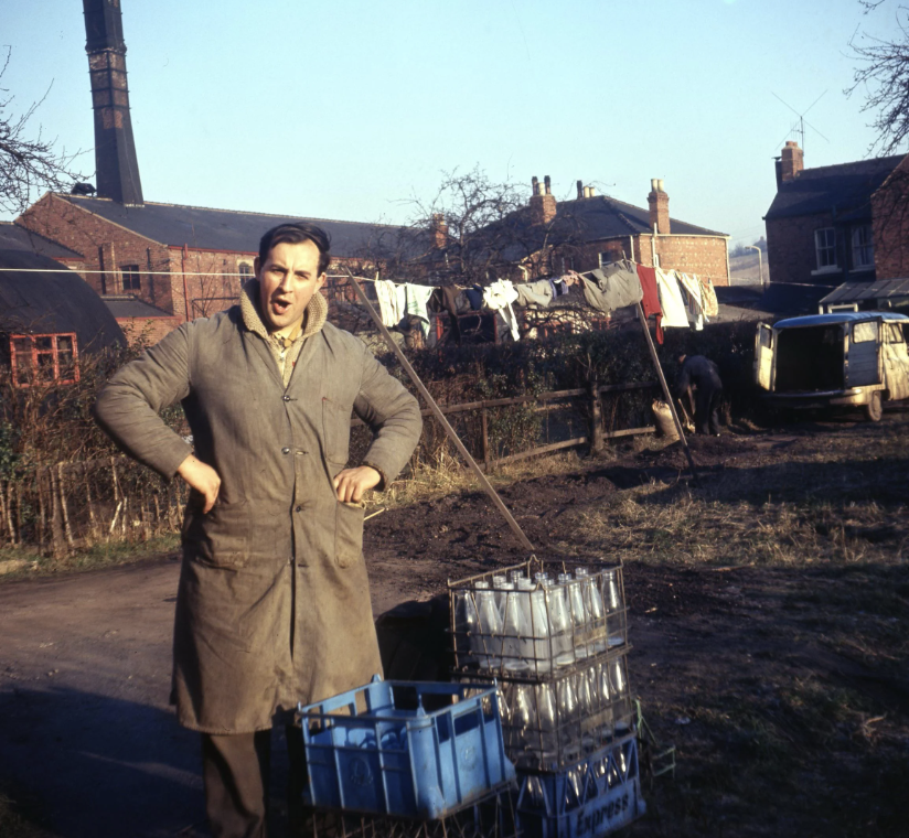 A person in a long coat stands with hands on hips on a rural street. Nearby, there are crates of bottles, a clothesline with hanging laundry, and a van. Brick buildings and a tall chimney are seen in the background under a clear sky.