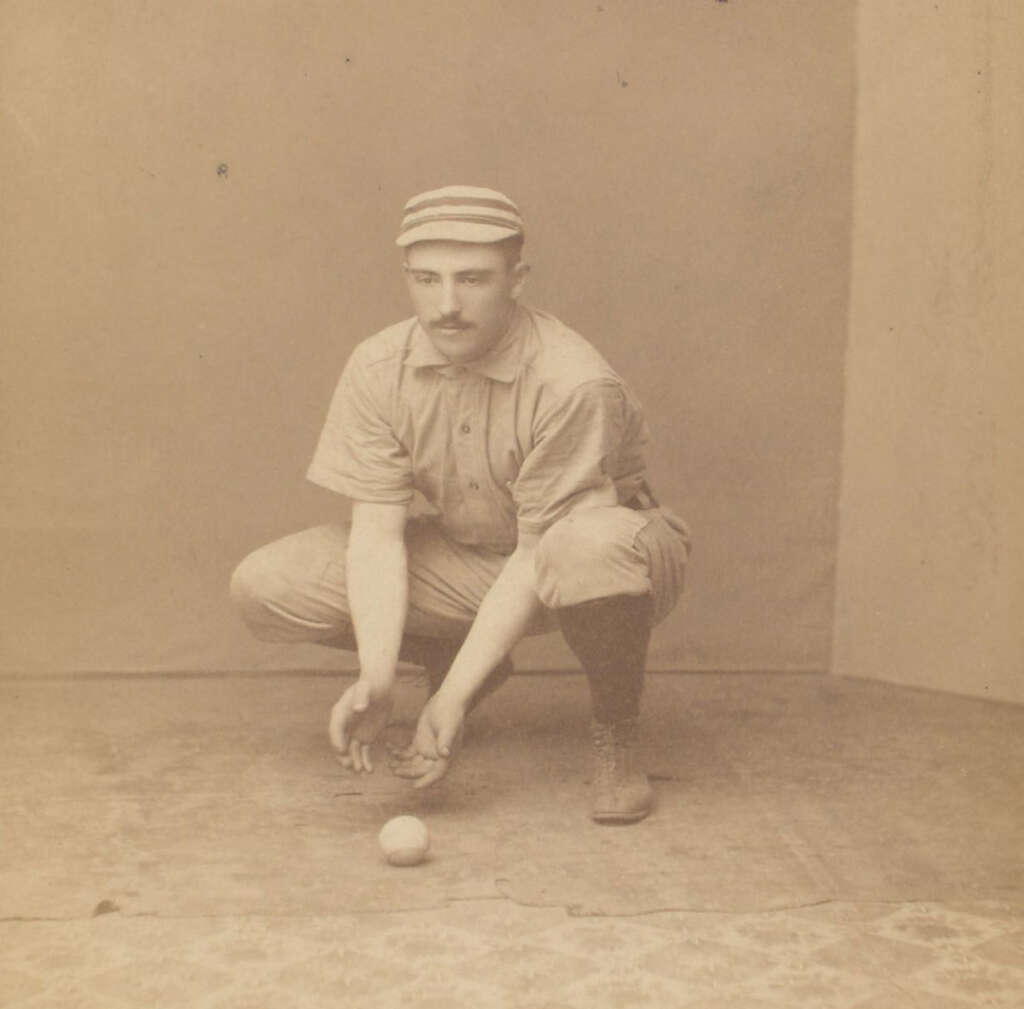 A vintage photograph of a baseball player in a striped cap, crouching with hands ready to catch a ball on the ground. He wears antique baseball attire, including a light-colored uniform and dark socks. The setting appears to be an indoor studio.