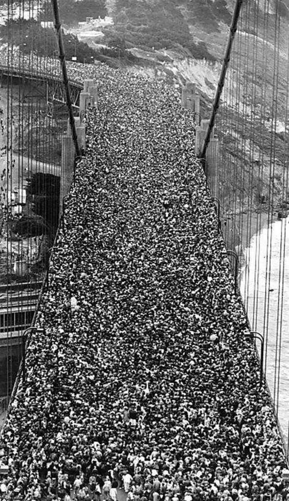 A historical black-and-white photo showing a massive crowd of people tightly packed and walking across a large suspension bridge. Their density covers the entire span of the bridge from end to end.