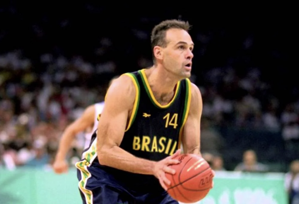 A basketball player in a Brazil jersey, numbered 14, is poised to shoot a basketball. He is focused, holding the ball with both hands. The background is blurred, with spectators visible.