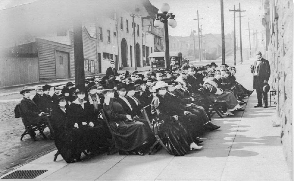 A historical black-and-white photo of a large group of people seated outdoors on a sidewalk. They are wearing coats, hats, and face masks. A man stands at the edge of the group. Buildings and a trolley are visible in the background.